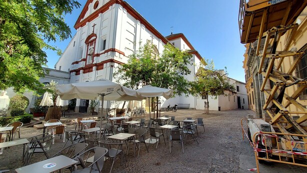 Ein Straßencafé mit Tischen und Stühlen, ein weißes Gebäude mit rotem Dach im Hintergrund und eine Baustelle rechts.