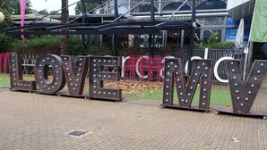 Ein großes Schild im Freien mit der Aufschrift "LOVE MY" in metallischen Buchstaben mit Beleuchtung, vor dem sich ein Gebäude mit Metallzaun und Grünflächen abhebt.