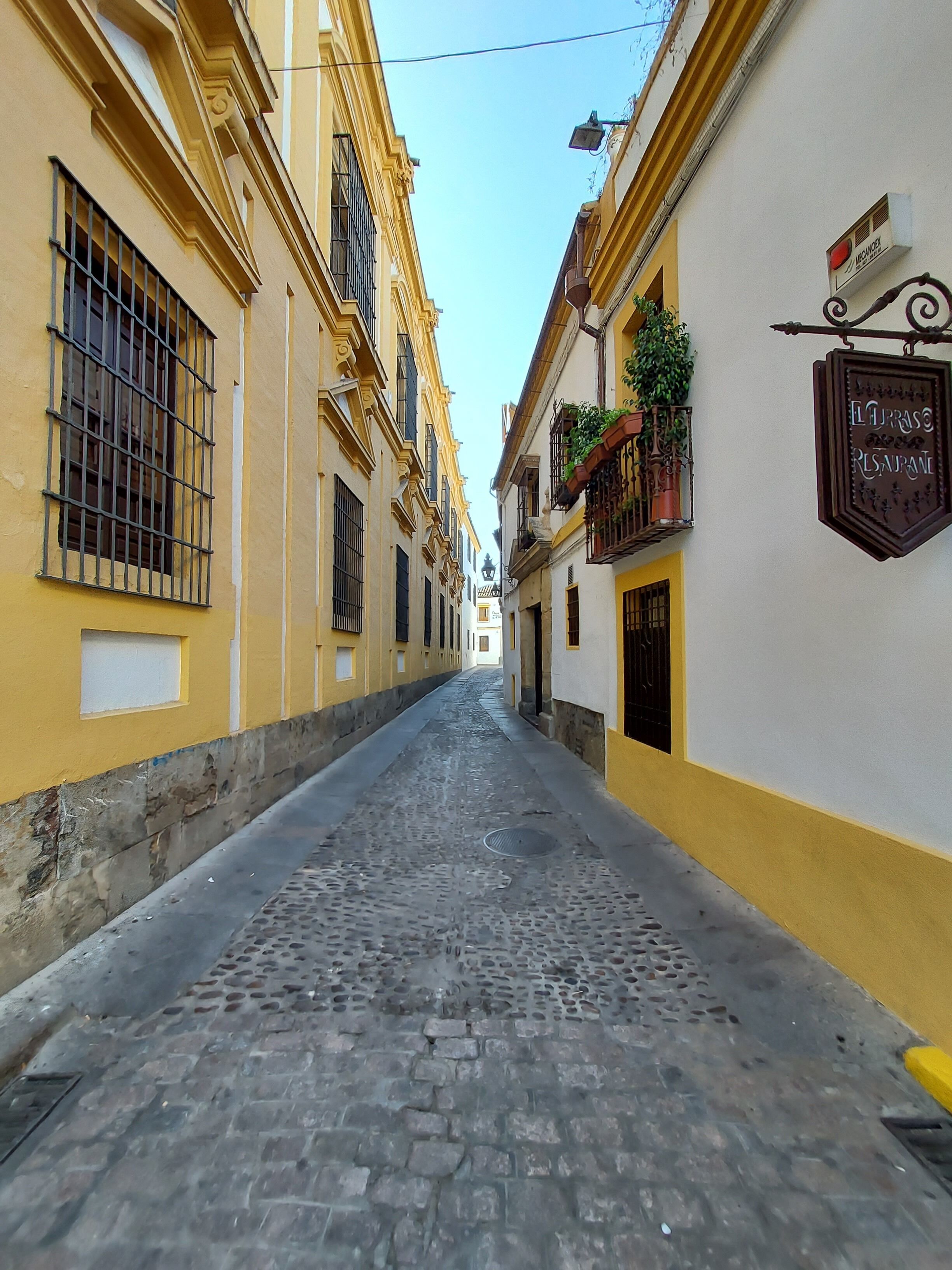 A narrow cobblestone street flanked by yellow buildings with barred windows and a sign for "El Prado Restaurant".