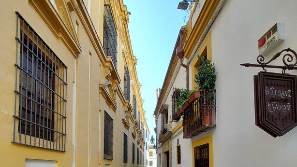 Une étroite rue pavée bordée de bâtiments jaunes aux fenêtres grillagées et sur laquelle on peut voir une enseigne indiquant le « Restaurant El Prado ».