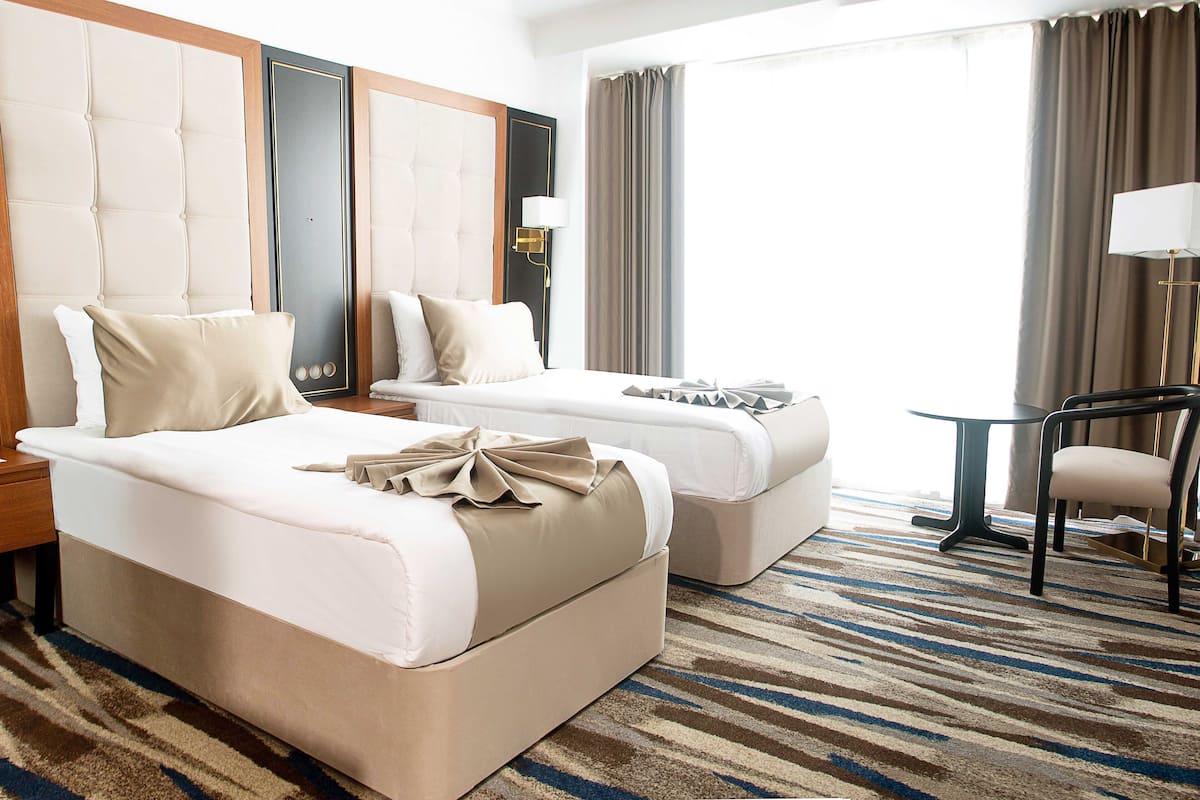 Quarto de hotel com duas camas, uma mesa pequena, uma cadeira e uma janela grande com cortinas.