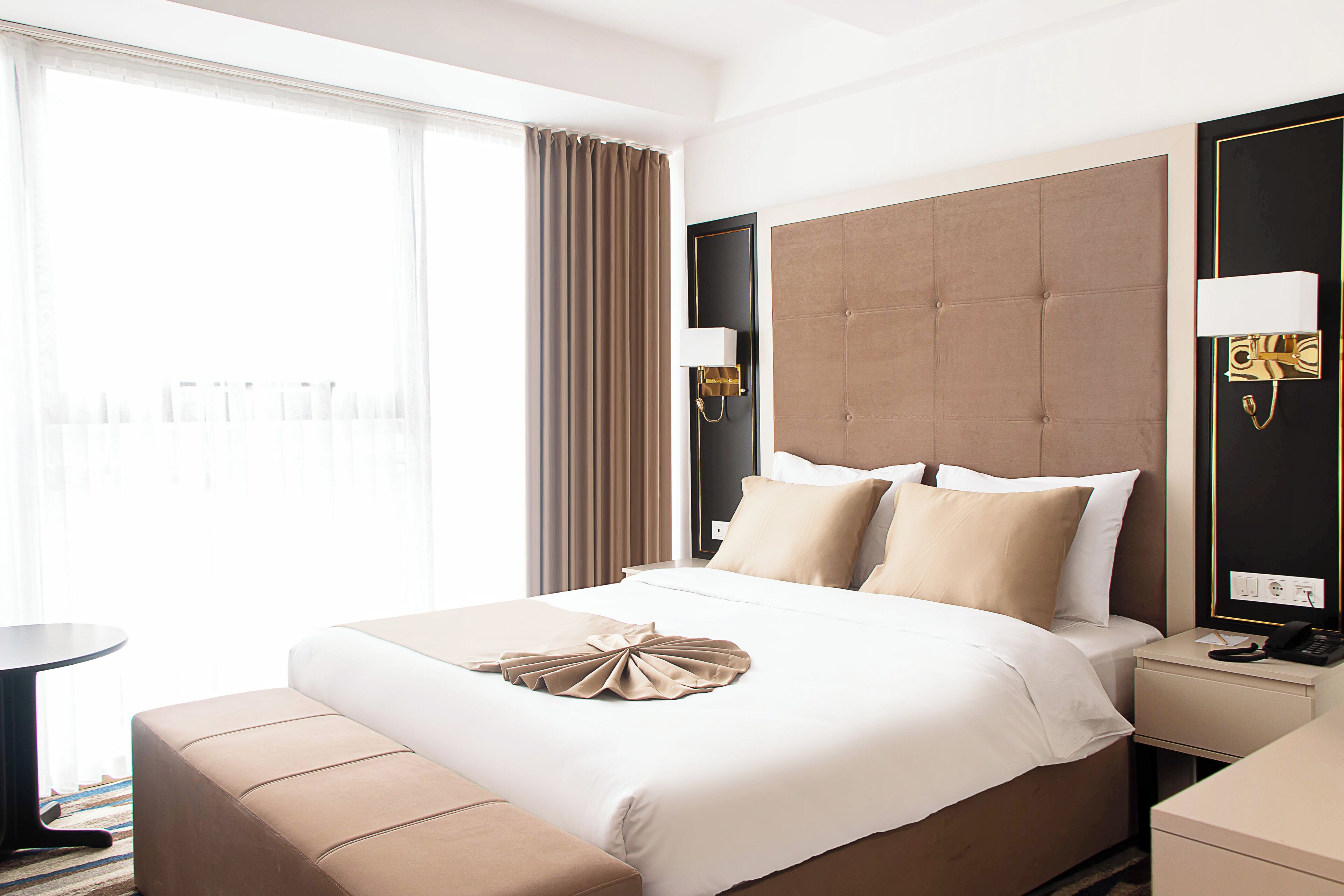 A hotel room with a bed, bedside table, lamp, and a view of the outside.