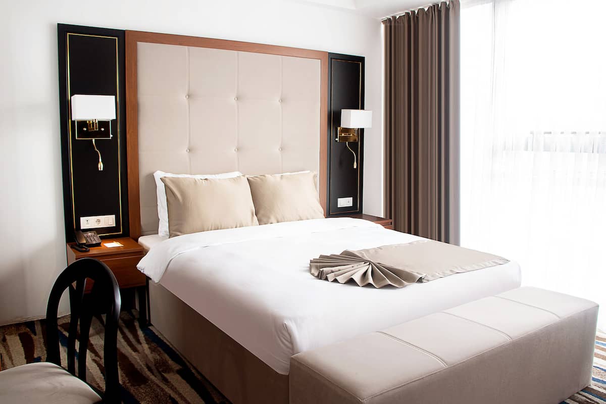 Quarto de hotel com uma cama grande, mesa de cabeceira com abajur, uma cadeira e vista para o exterior pela janela.