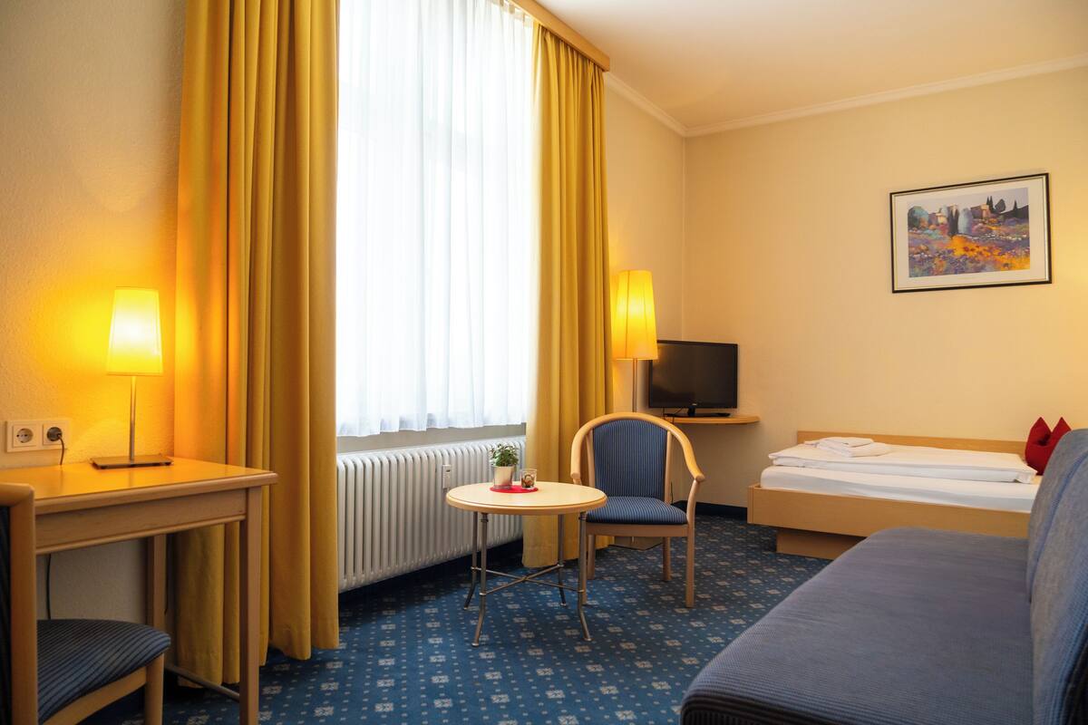 Una habitación de hotel con cama, escritorio con lámpara, una silla, una mesita y televisión.