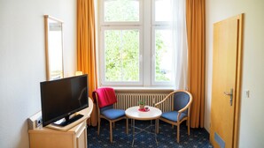 A hotel room with a television, two chairs, a small table, a window with curtains, and a radiator. - Bischofshof Braustuben (Regensburg)