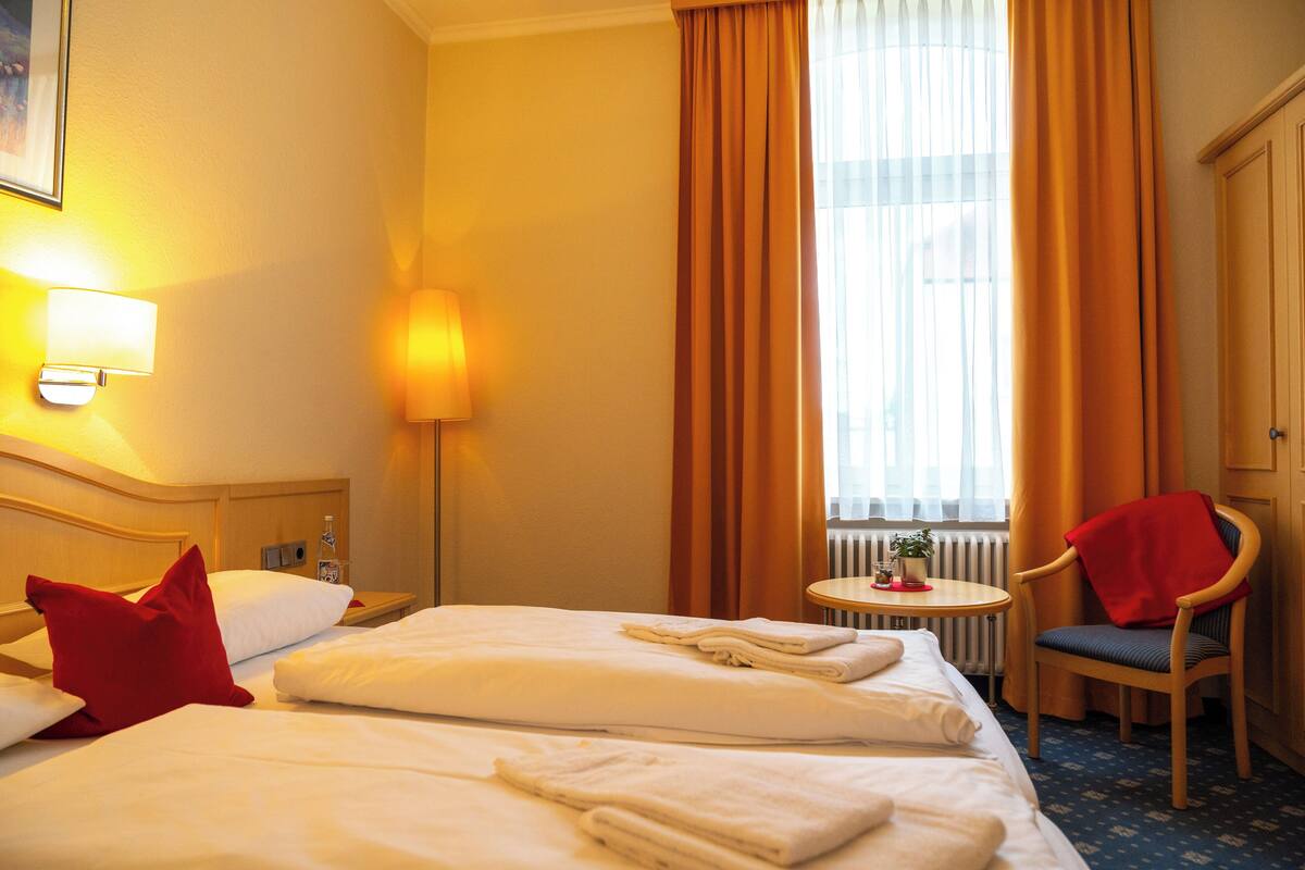 Habitación de hotel con dos camas, una silla, una mesa y un ventanal con cortinas.