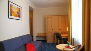 A hotel room with a blue sofa, a small table with a plant, a wooden cabinet, and a framed picture on the wall. - Bischofshof Braustuben (Regensburg)