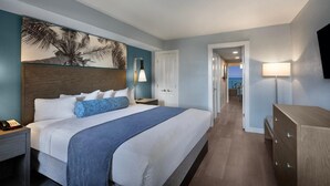 Ein Hotelzimmer mit einem großen Bett, einem Nachttisch mit Telefon, einer Schreibtischlampe, einem Schminktisch und einem Palmenbaum-Gemälde.