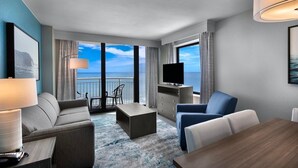 Ein modernes Hotelzimmer mit Balkon, einer Couch, einem Sessel, einem Fernseher und einem Esstisch.