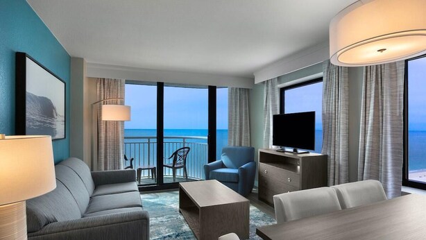 Ein modernes Hotelzimmer mit Balkon, einer Couch, einem Fernseher und einem Esstisch.