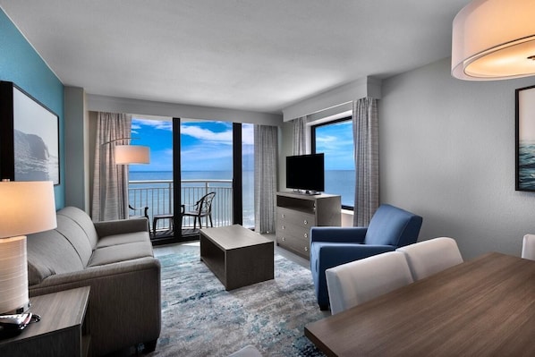 Ein modernes Hotelzimmer mit Balkon, Fernseher, Sofa, Sessel und Esstisch.
