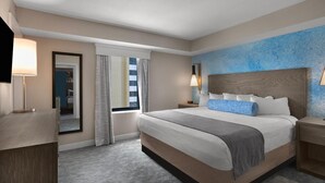 Quarto de hotel com uma cama grande, uma mesa de cabeceira, uma luminária, um espelho e uma janela com cortinas.