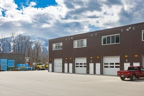 A large industrial building with multiple garage doors, a red pickup truck parked in front, and a mountainous background. - The Stellar Condo - Cozy 2 Bedroom with Hot Tub (Revelstoke)