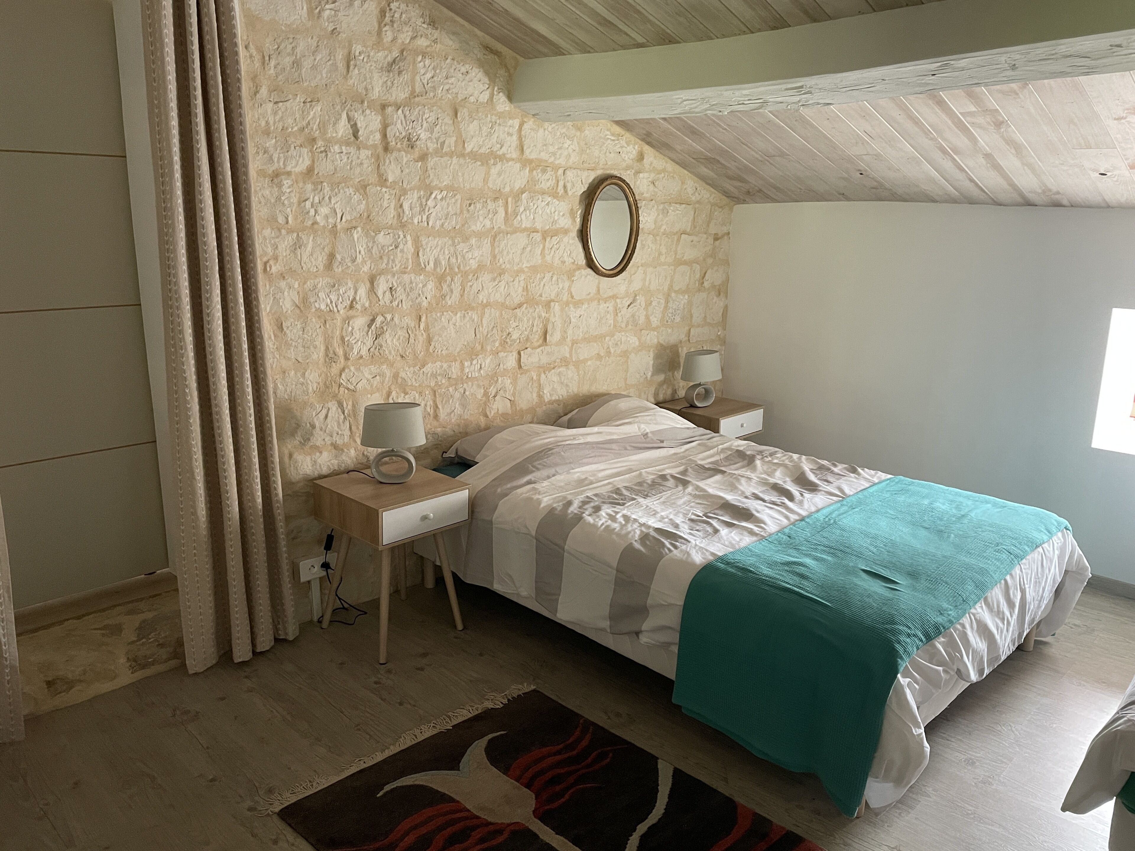 A bedroom with a stone wall, a bed with a teal blanket, two bedside tables with lamps, and a window.