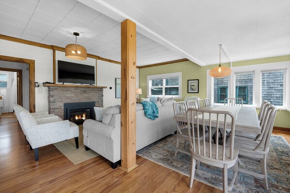 A living room with a fireplace, a dining area with a table and chairs, and a television mounted on the wall. - SELAH BY THE SEA - Beachfront! (Seaside)
