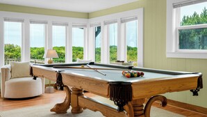 Una mesa de billar con taco, bolas y lámpara en una habitación con amplios ventanales y vista a zonas verdes.
