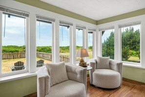 A room with large windows, two armchairs, a small table with a lamp, and a view of a fenced outdoor area. - SELAH BY THE SEA - Beachfront! (Seaside)