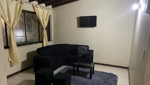 Una sala de estar con un sofá negro, dos otomanas negras, una mesita de centro pequeña y un tapete azul. Hay un televisor montado en la pared y una ventana con cortinas.