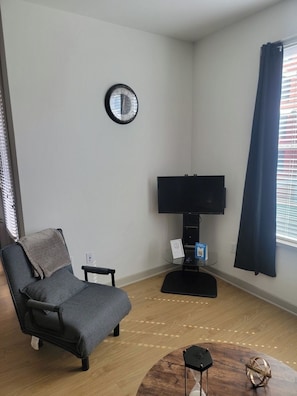 Ett vardagsrum med en grå fåtölj, en platt-TV på ett TV-bord, en klocka på väggen och ett fönster med gardiner.