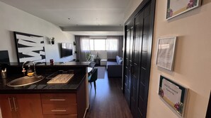 Una cocina moderna con isla integrada, un comedor con mesa y sillas, una sala de estar con sofá y televisor, y un pasillo con una puerta.