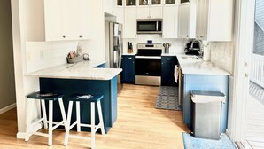 Cozinha moderna com armários brancos, eletrodomésticos de aço inoxidável, uma ilha central com banquetas e vista para a sala de estar adjacente.