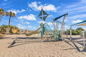 A playground with a slide and climbing structures, surrounded by sand and palm trees.