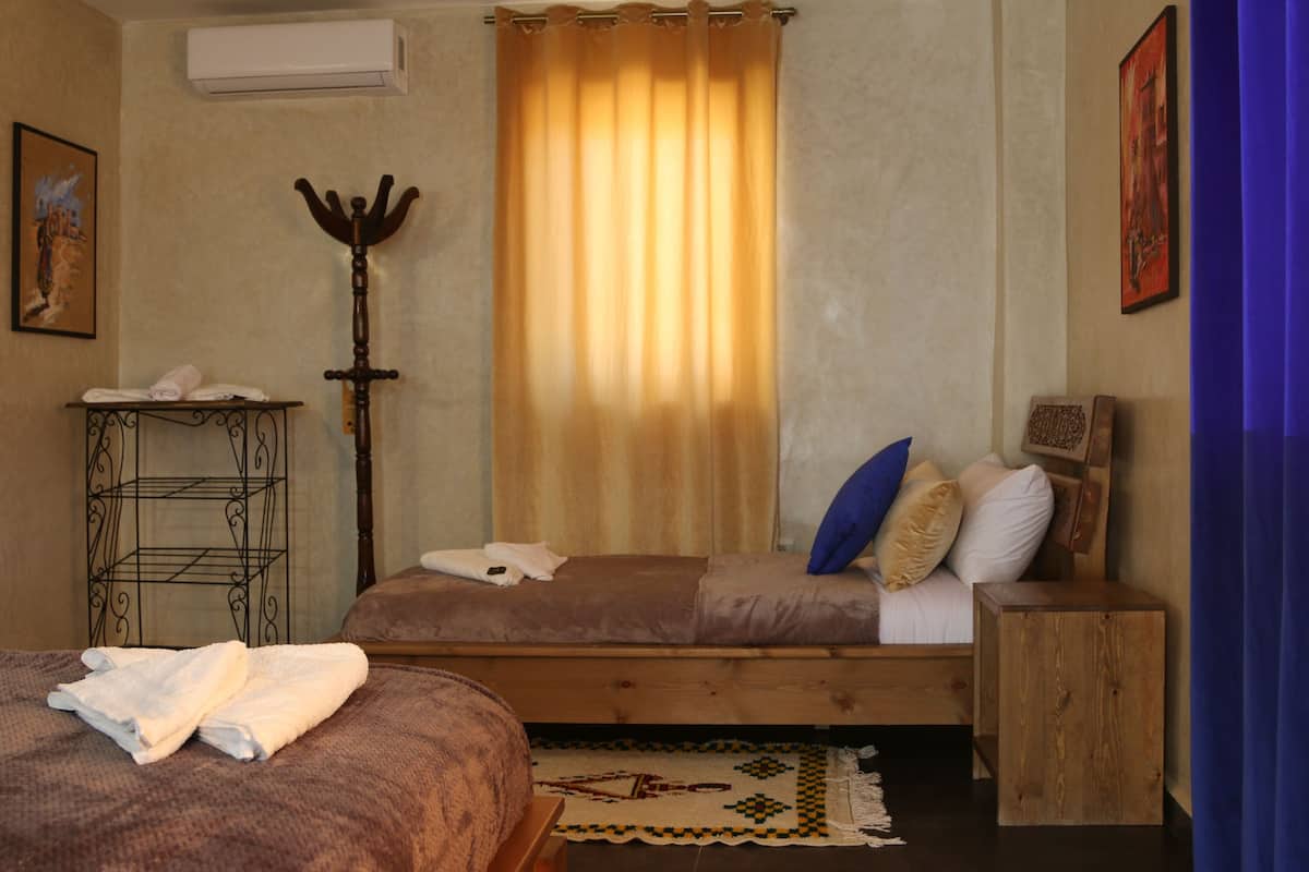 Een kamer met twee bedden, een houten bankje, een tafeltje met een handdoek, een decoratieve staf en een kamerkleed met een patroon.