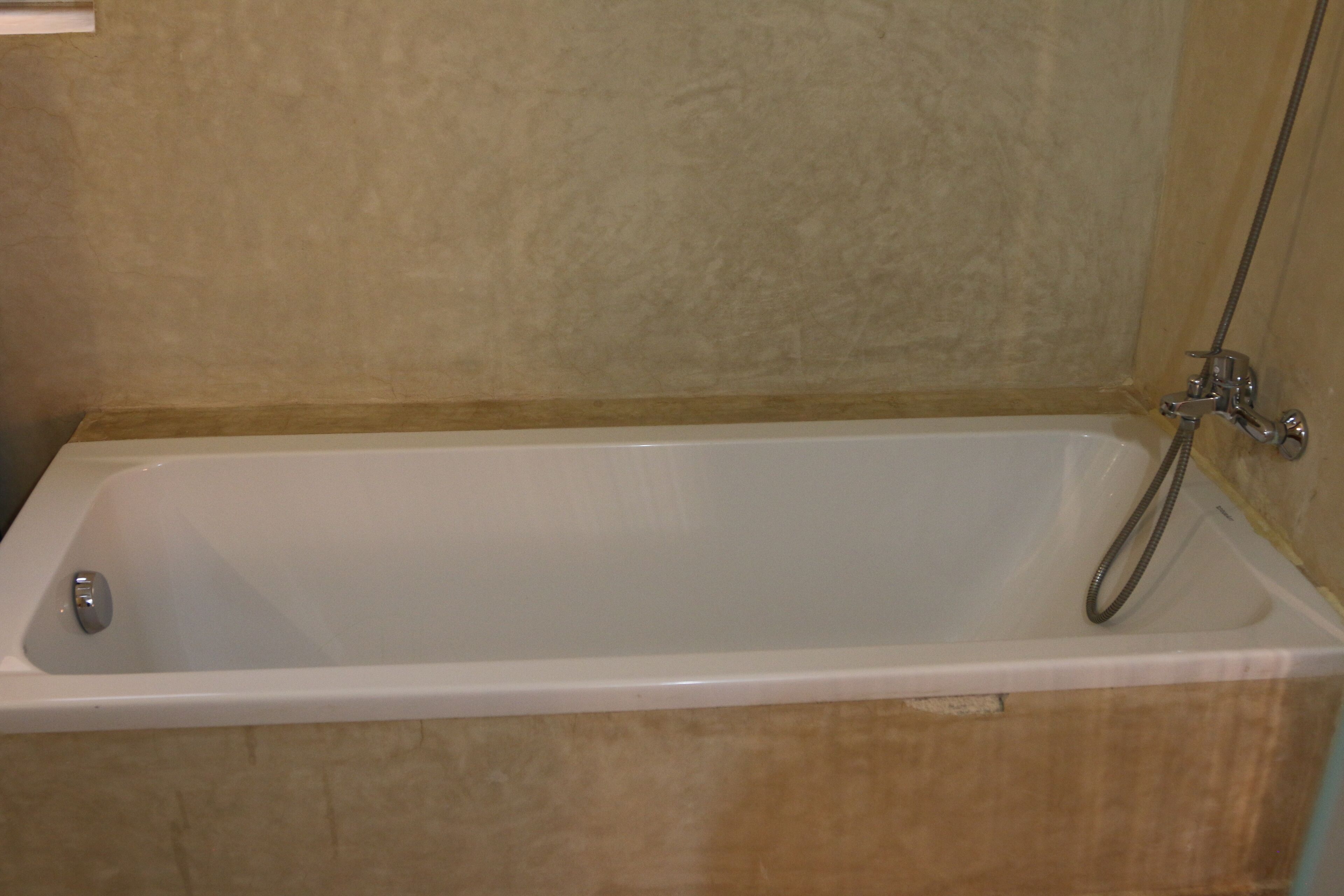 A standard rectangular bathtub with a shower head and faucet, set against a textured beige wall.