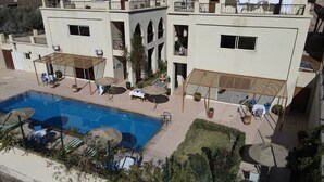Un espace piscine doté de chaises longues et de parasols, entouré d’un bâtiment avec des balcons et une terrasse couverte.