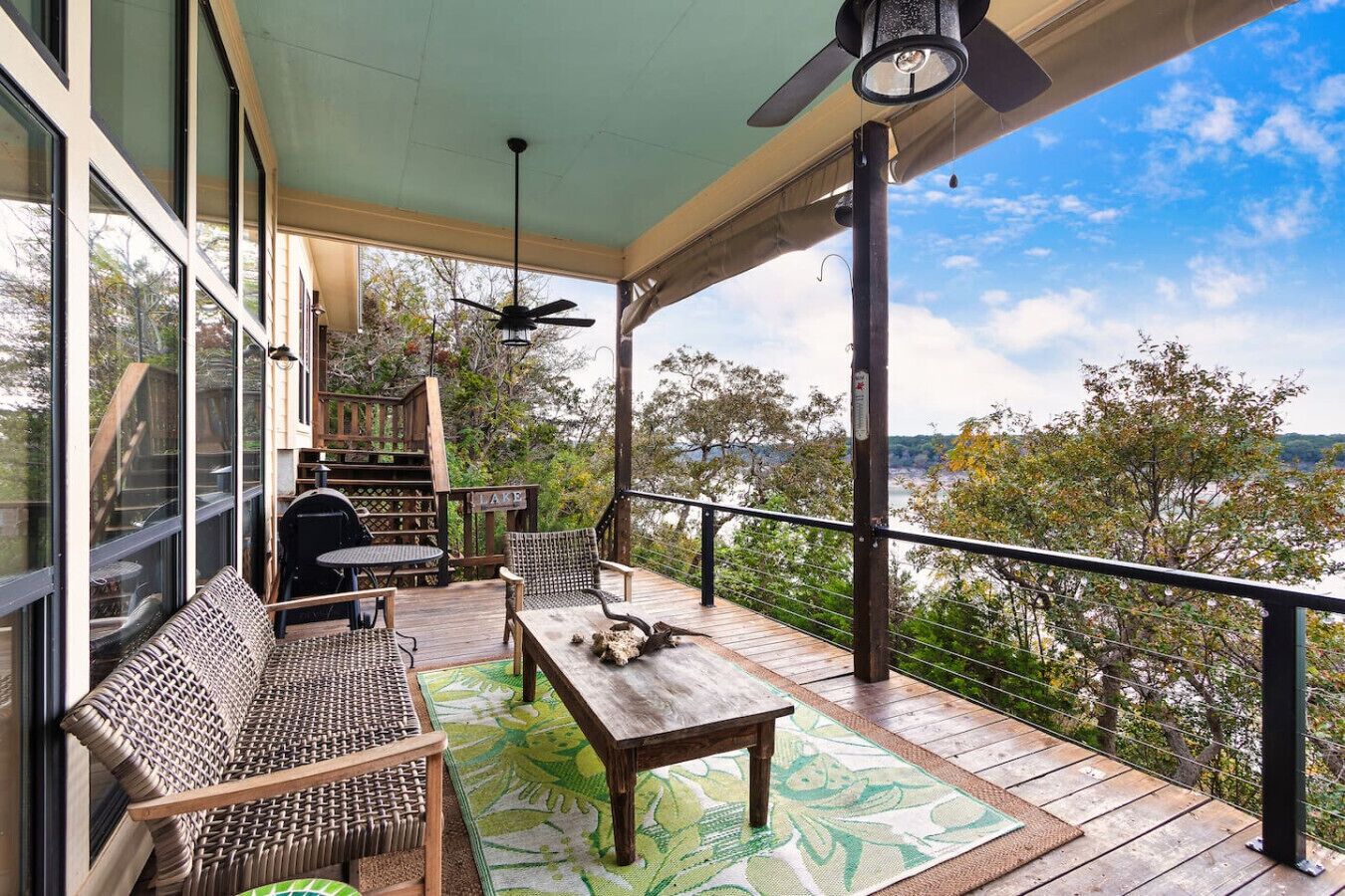 Une terrasse extérieure couverte, équipée d’une table et de chaises, d’un barbecue, et offrant une vue sur les arbres et un lac.