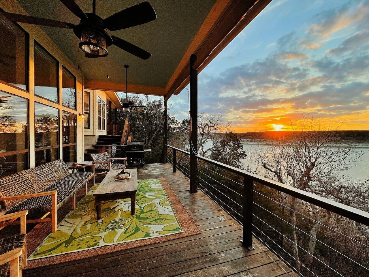 Une terrasse en bois avec une table et des chaises, donnant sur une étendue d’eau au coucher du soleil.