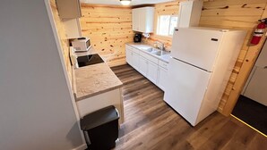 A compact kitchen with wooden walls, white cabinets, a sink, a microwave, a refrigerator, and a trash bin.