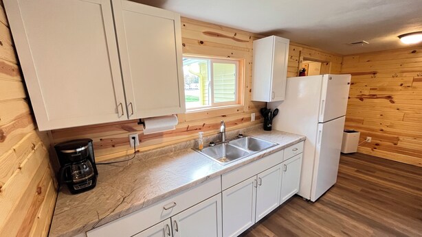 A kitchen with wooden walls, white cabinets, a double sink, a coffee maker, and a refrigerator.