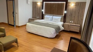 Una habitación de hotel moderna con una cama grande, mesitas de noche, cabecero de madera y una zona de estar con una silla y reposapiés.