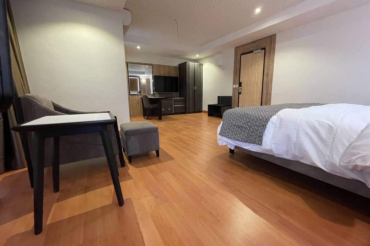 Quarto de hotel moderno com piso de madeira, cama com manta estampada, escrivaninha com cadeira, um pequeno banquinho, TV e cozinha compacta ao fundo.