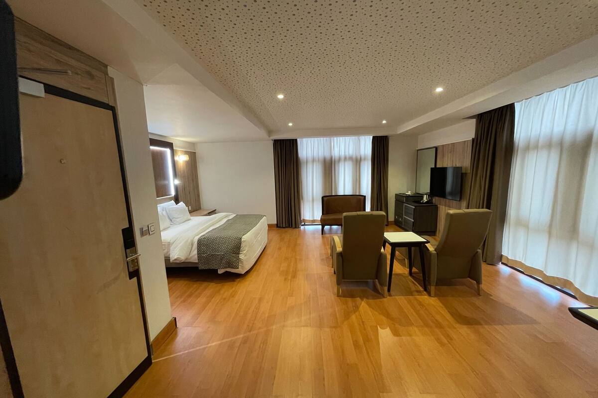 Quarto de hotel com uma cama, uma escrivaninha com cadeira, uma televisão e uma janela grande com cortinas.