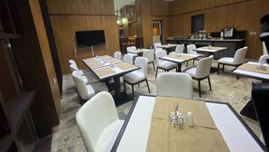 Un comedor moderno de hotel con varias mesas y sillas, una cocina al fondo y un televisor de pantalla plana montado en la pared.