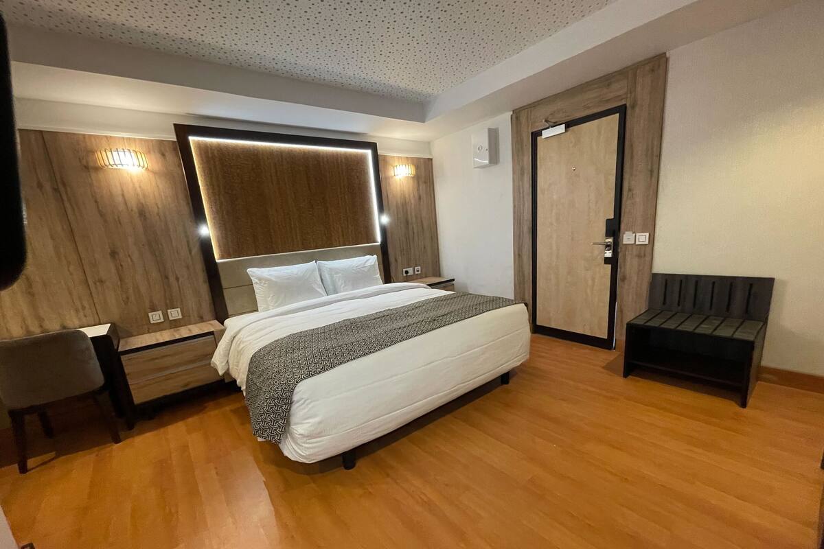 Quarto de hotel moderno com uma cama grande, cabeceira de madeira, um banco e um guarda-roupa.