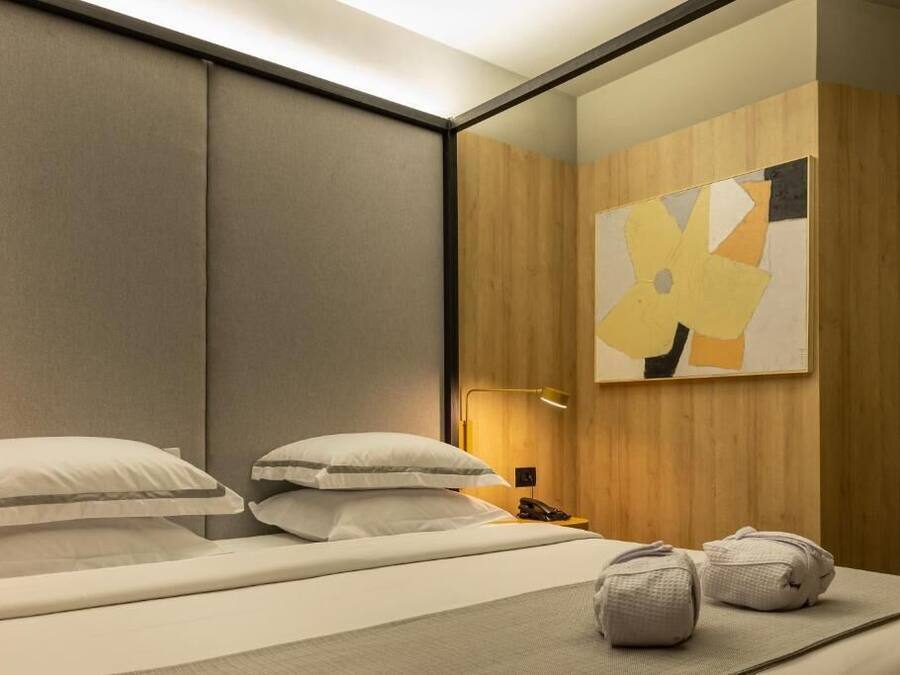 Habitación de hotel con cama, almohadas, mesita de noche con lámpara y una obra de arte en la pared.