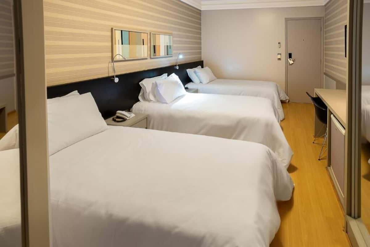A hotel room with three single beds, a wooden headboard, a bedside table with a lamp, and a small desk with a chair.
