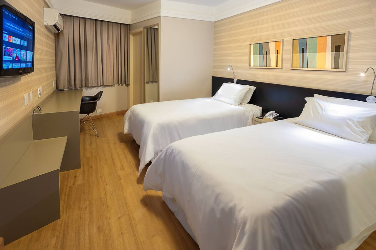 A hotel room with two beds, a desk, a chair, a TV, and a wall-mounted air conditioner.