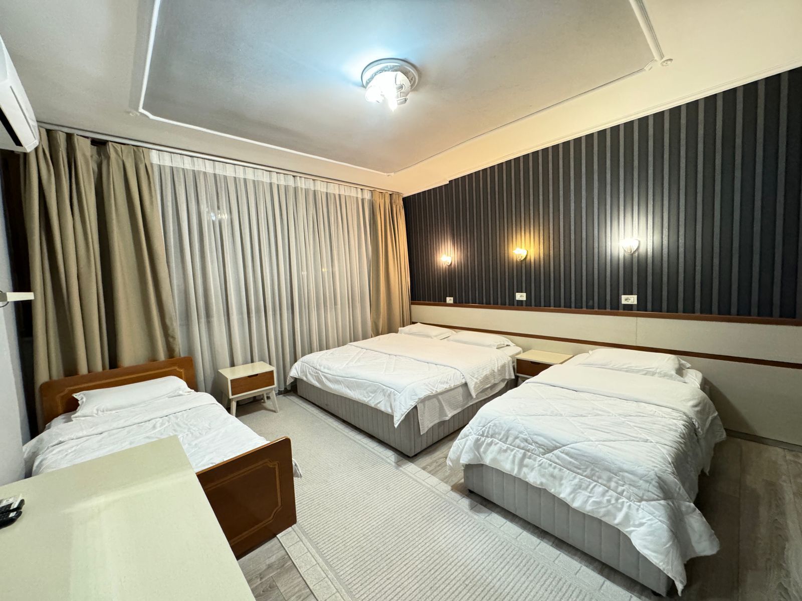Habitación de hotel con tres camas, una mesita de noche pequeña, un espejo grande y una lámpara en el techo.