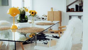 Een moderne eethoek met een glazen tafel, witte stoelen, een gele lamp en een vaas met bloemen.