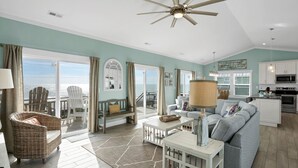 A spacious living room with a large sofa, a dining area with chairs, a kitchen with modern appliances, and a balcony with a view of the ocean. - Wright By The Sea - premier 4 bedroom oceanfront home (North Topsail Beach)