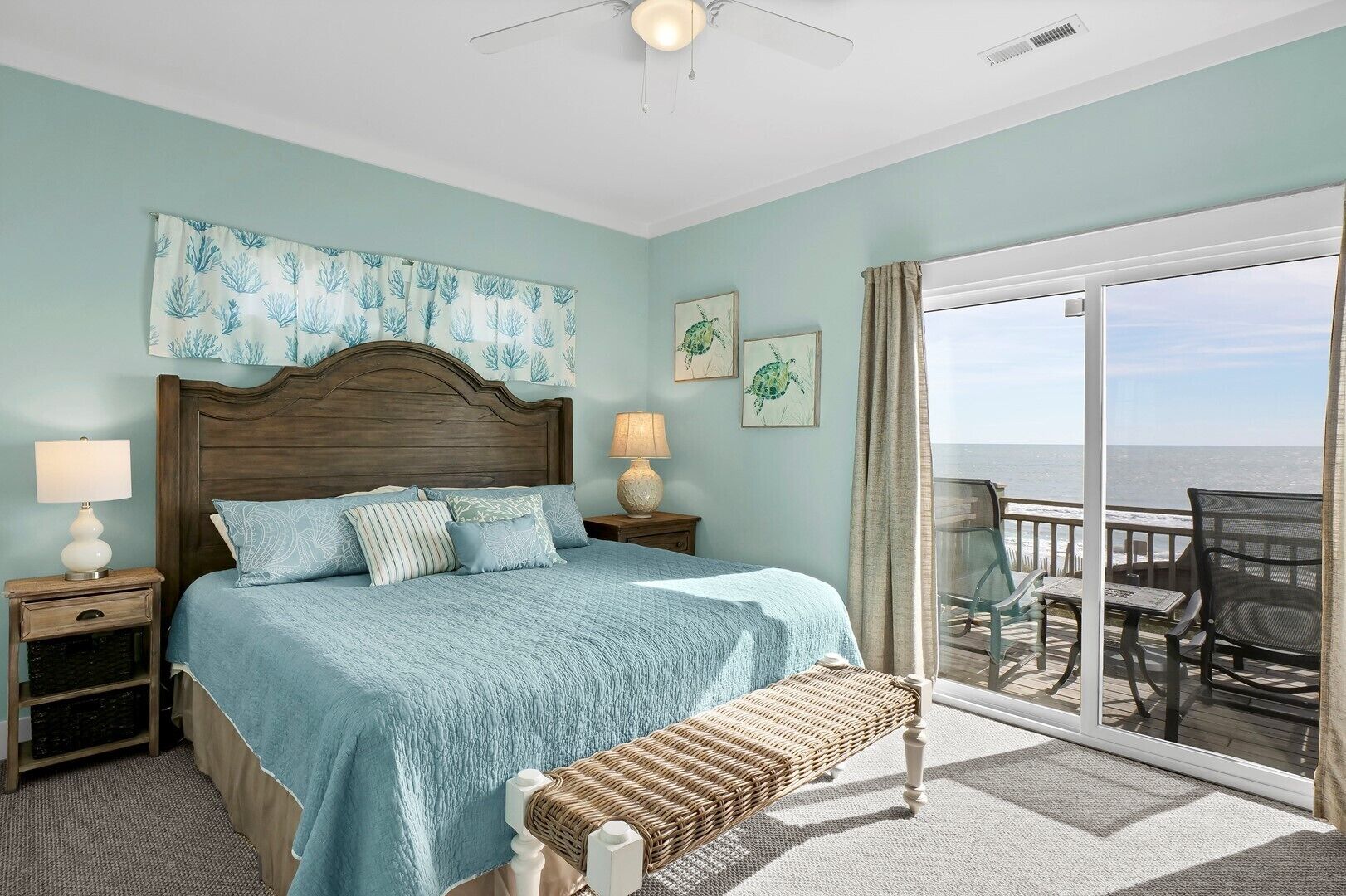 A bedroom with a large bed, two bedside tables with lamps, a bench, and a view of the ocean through a sliding glass door.
