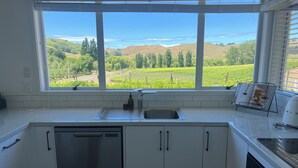 A kitchen with white cabinets, a dishwasher, a sink, and a window with a view of green fields and hills. - Private setting with vineyard outlook yet only moments from the Village.  (Havelock North)