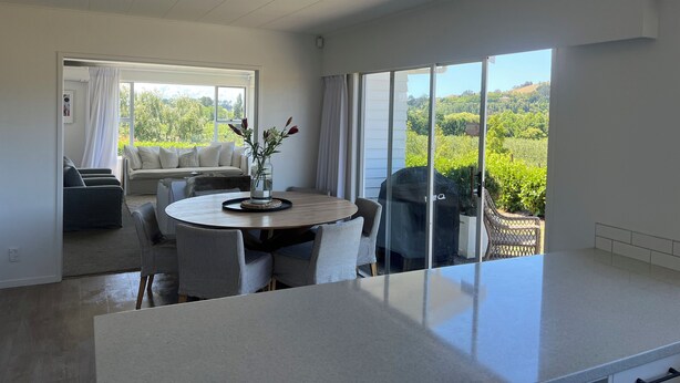 A modern living room with a round dining table, a sofa, and a large sliding glass door leading to a balcony with a view of greenery. - Private setting with vineyard outlook yet only moments from the Village.  (Havelock North)