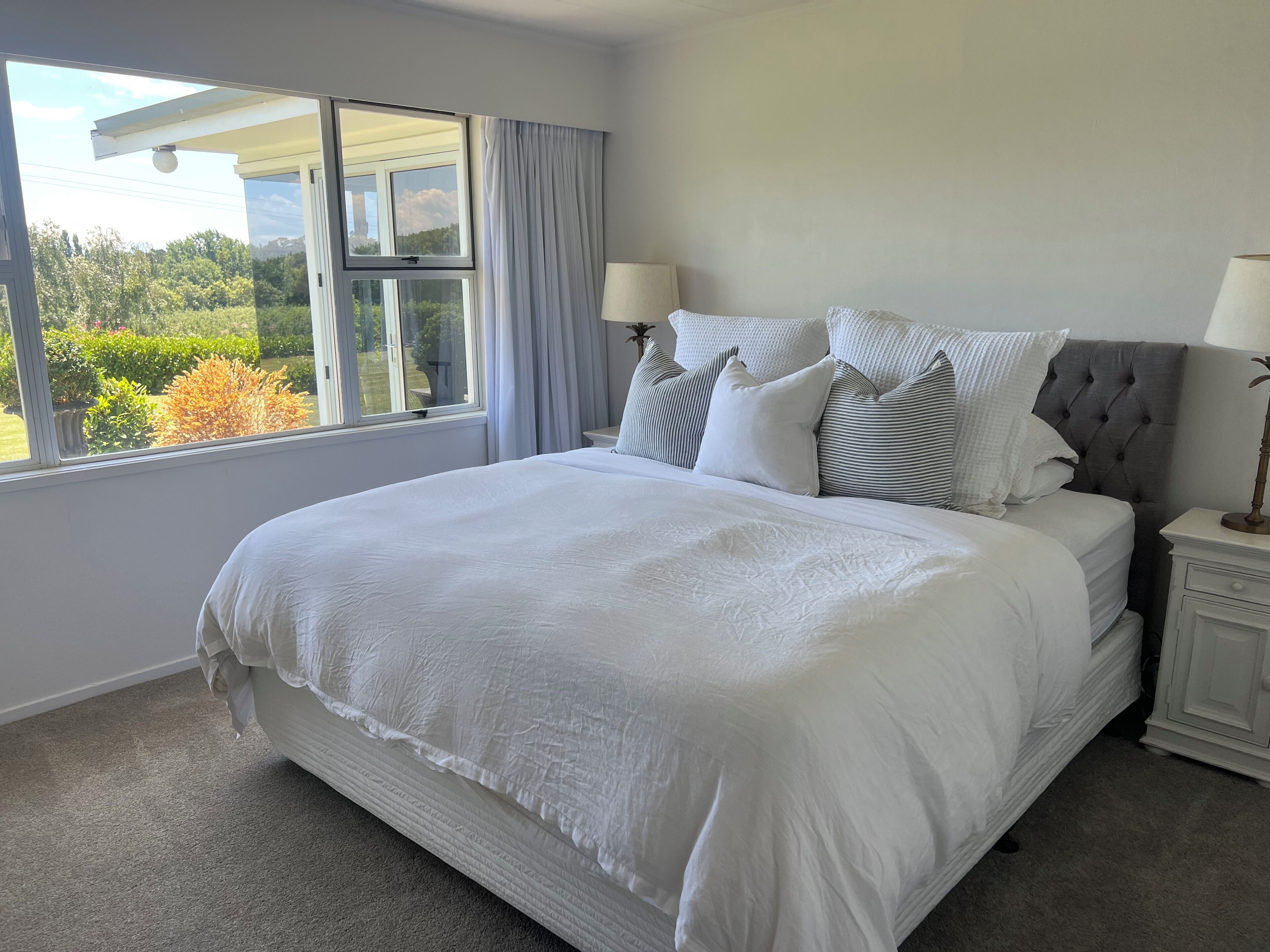 A neatly made bed with white linens, a grey upholstered headboard, and multiple pillows. There is a window with a view of greenery outside.