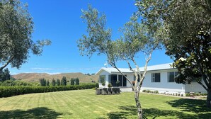 A white house with a covered patio, surrounded by a well-maintained lawn and trees. - Private setting with vineyard outlook yet only moments from the Village.  (Havelock North)