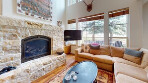 A stone fireplace, a flat-screen TV, a beige sofa, and a coffee table with three glasses. - Central Park 26- Luxury Tri-Level in Great Location Close to River Run (Ketchum)